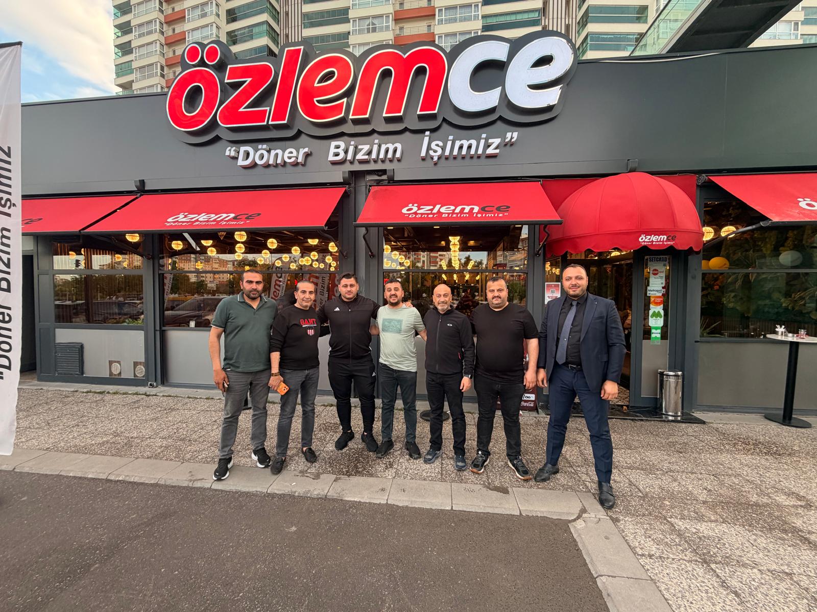 Seven men pose together in front of an Özlemce restaurant with red awnings and a bold red-and-white sign in a city setting, smiling at the camera.
